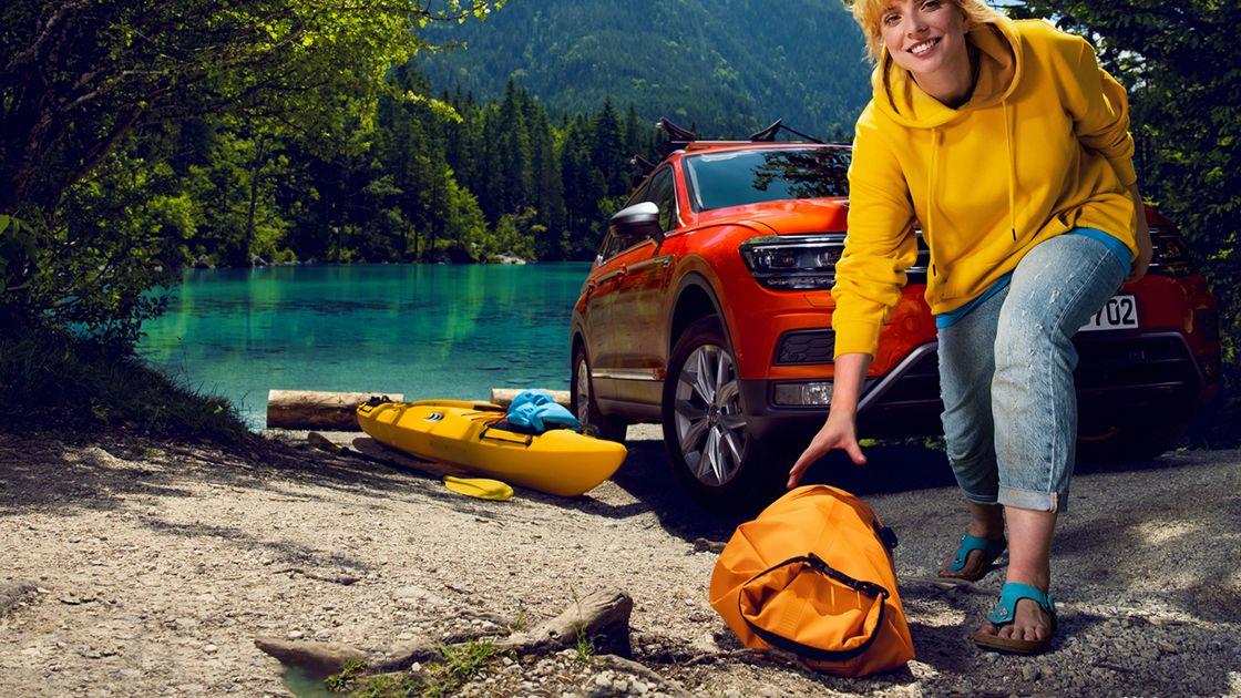 A brightly and casually dressed lady picking up her yellow dive bag in front of a red Volkswagen SUV by a lake.