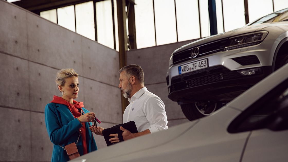 A Volkswagen Owner handing over the car key to a Certified Volkswagen Staff in a service workshop behind a white Volkswagen vehicle.