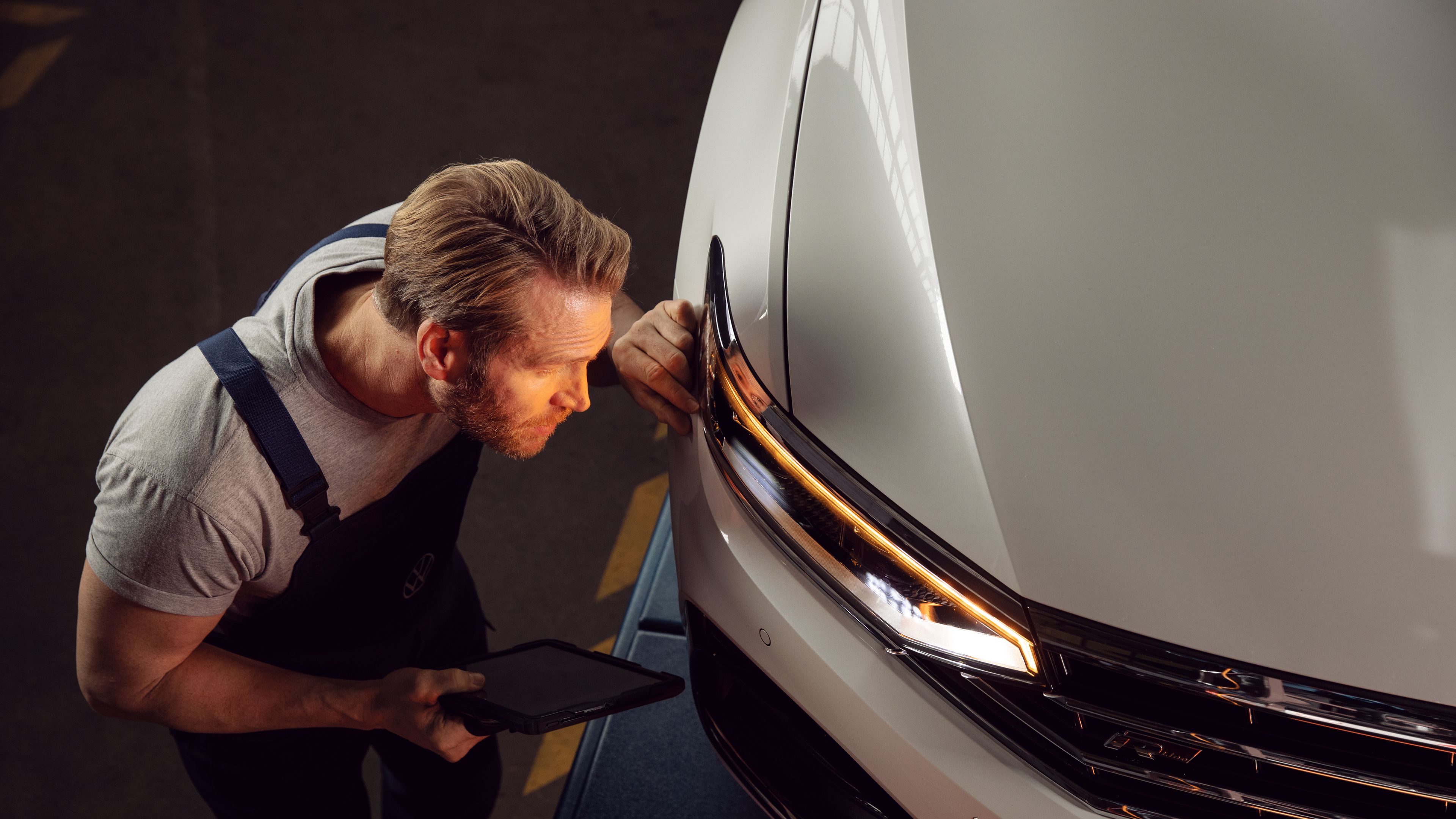 A certified Volkswagen Staff holding a smart tablet carefully checking the lit headlamps of a white Volkswagen vehicle for potential defects.