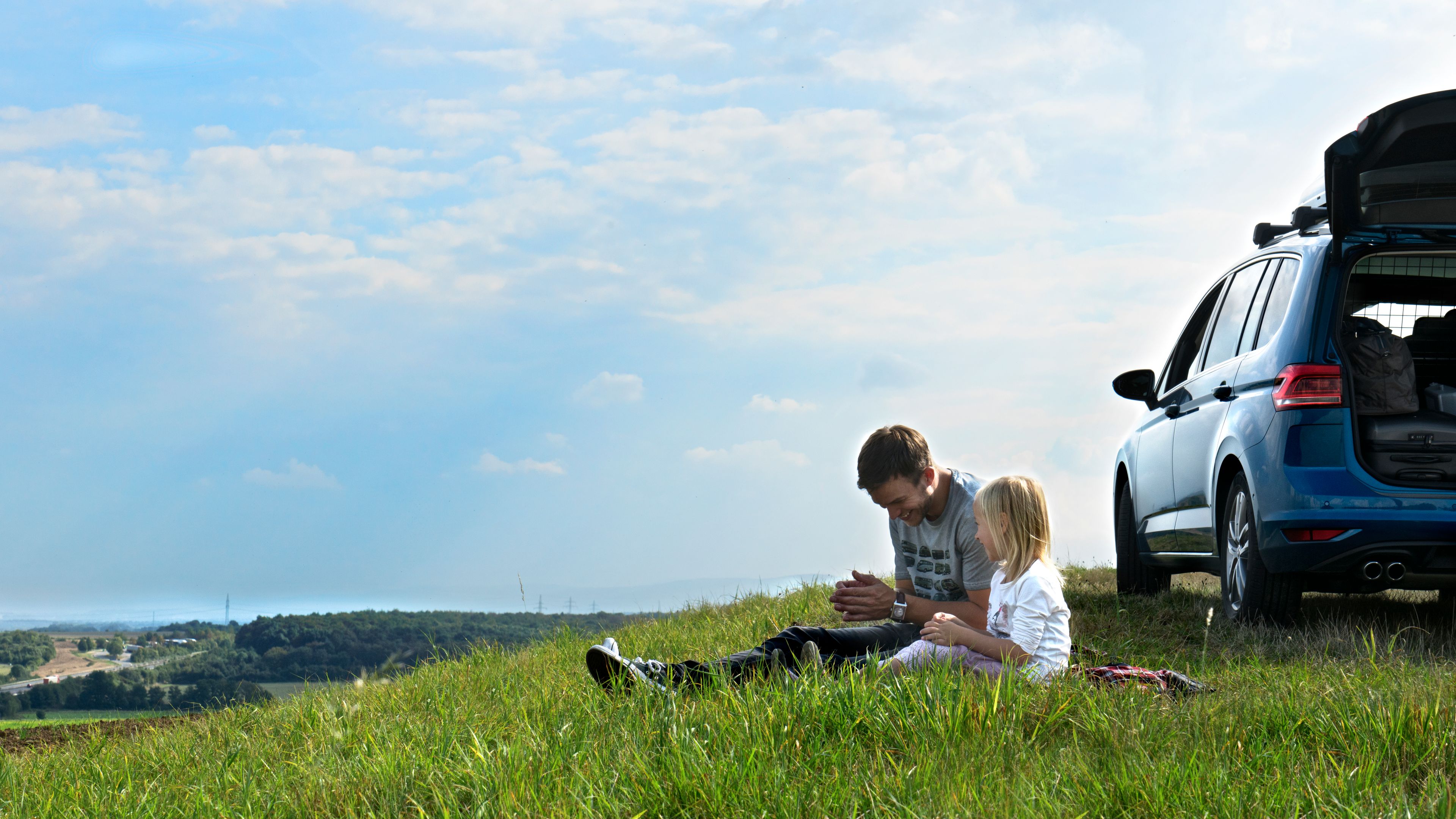 With clear blue skies, a father and daughter sit atop lush greenery with their blue Volkswagen behind them