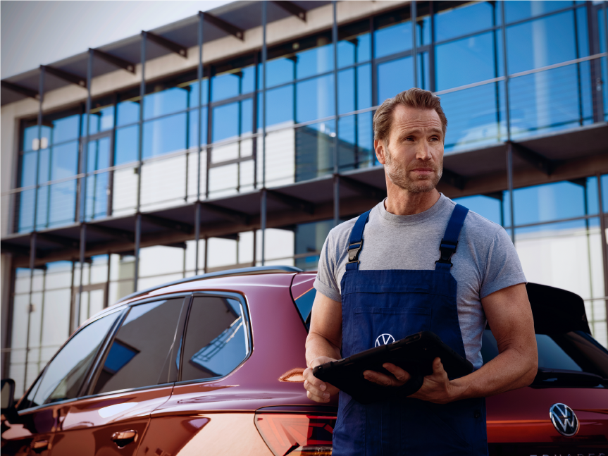 A man holding a tablet standing behind a red Volkswagen red car at an outdoor setting.