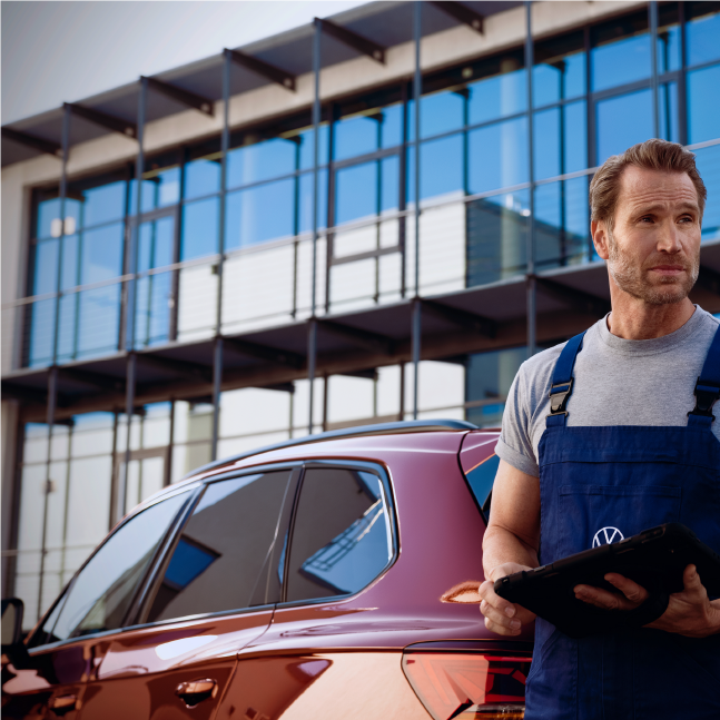 A man holding a tablet standing behind a red Volkswagen red car at an outdoor setting.