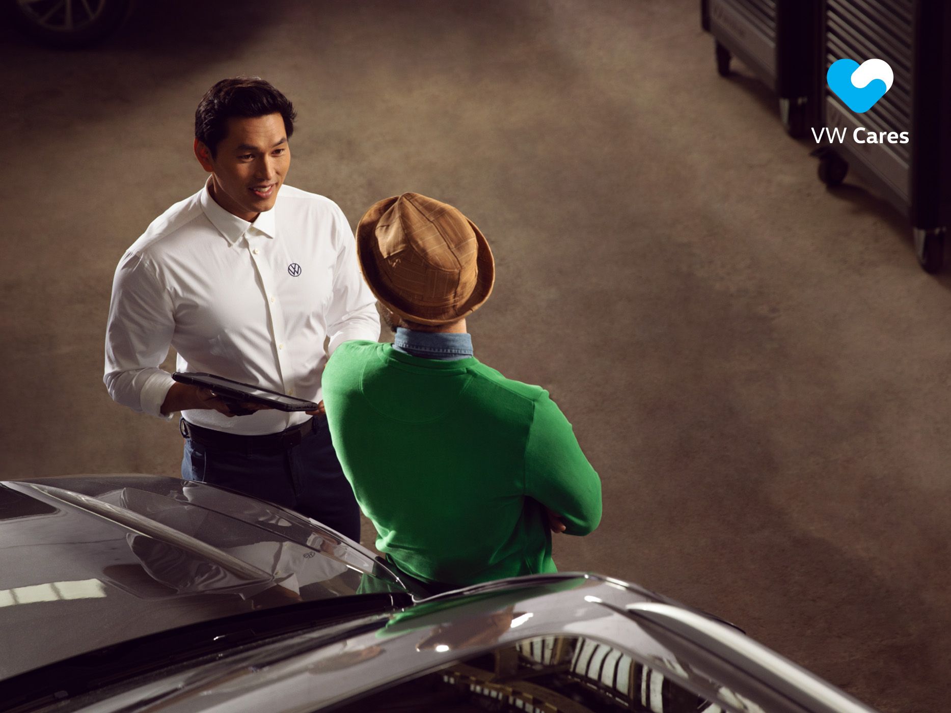 A man dressed in earth toned colour is seen having a service dialogue with a Volkswagen Service Advisor in a workshop setting.