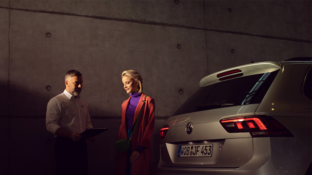 A Certified Volkswagen Staff providing consulting a Volkswagen Owner at a service workshop next to a silver Volkswagen SUV.