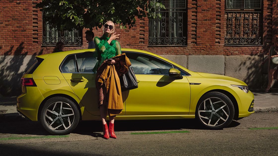 A chic looking lady wearing a pair of sunglasses while holding a handbag is seen standing beside a yellow Volkswagen Golf at a outdoor setting.