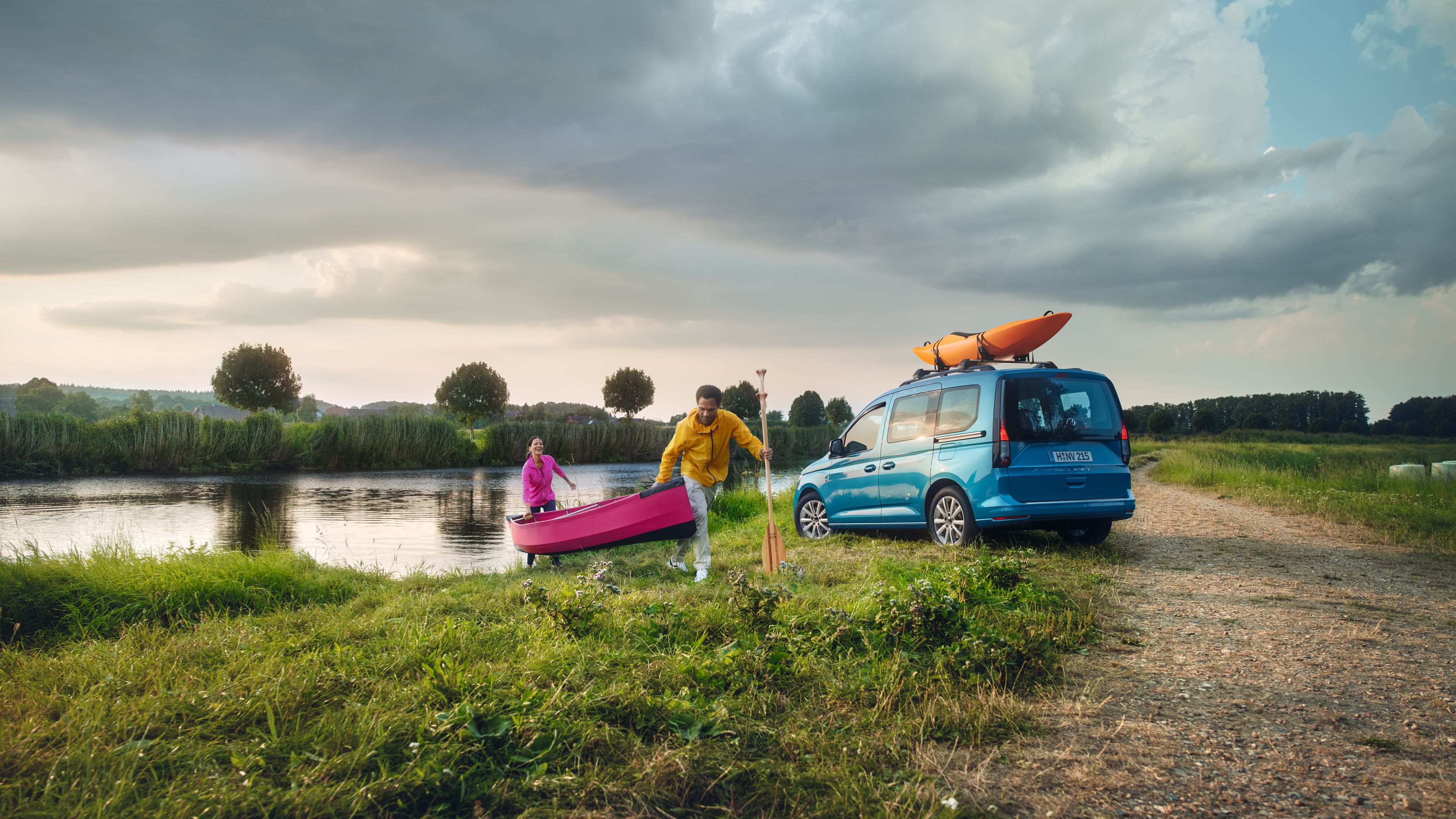 A couple dressed in outdoor-ready outfits carrying a pink coloured boat out from a lake beside a parked blue coloured light Volkswagen commercial vehicle at an outdoor setting.