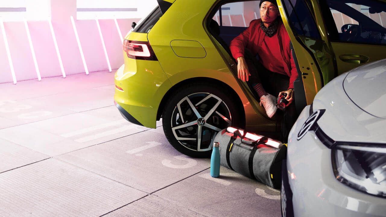 A model dressed in a stylish red outfit sitting at the backseat of a yellow Volkswagen vehicle alongside Genuine Volkswagen Merchandises such as the flask and barrel bag during a photo shooting session.