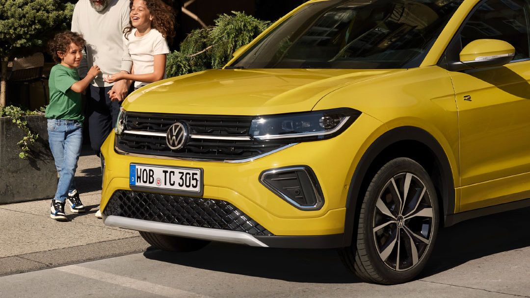 A family is seen walking beside a parked yellow Volkswagen T-Cross at an urban outdoor setting.