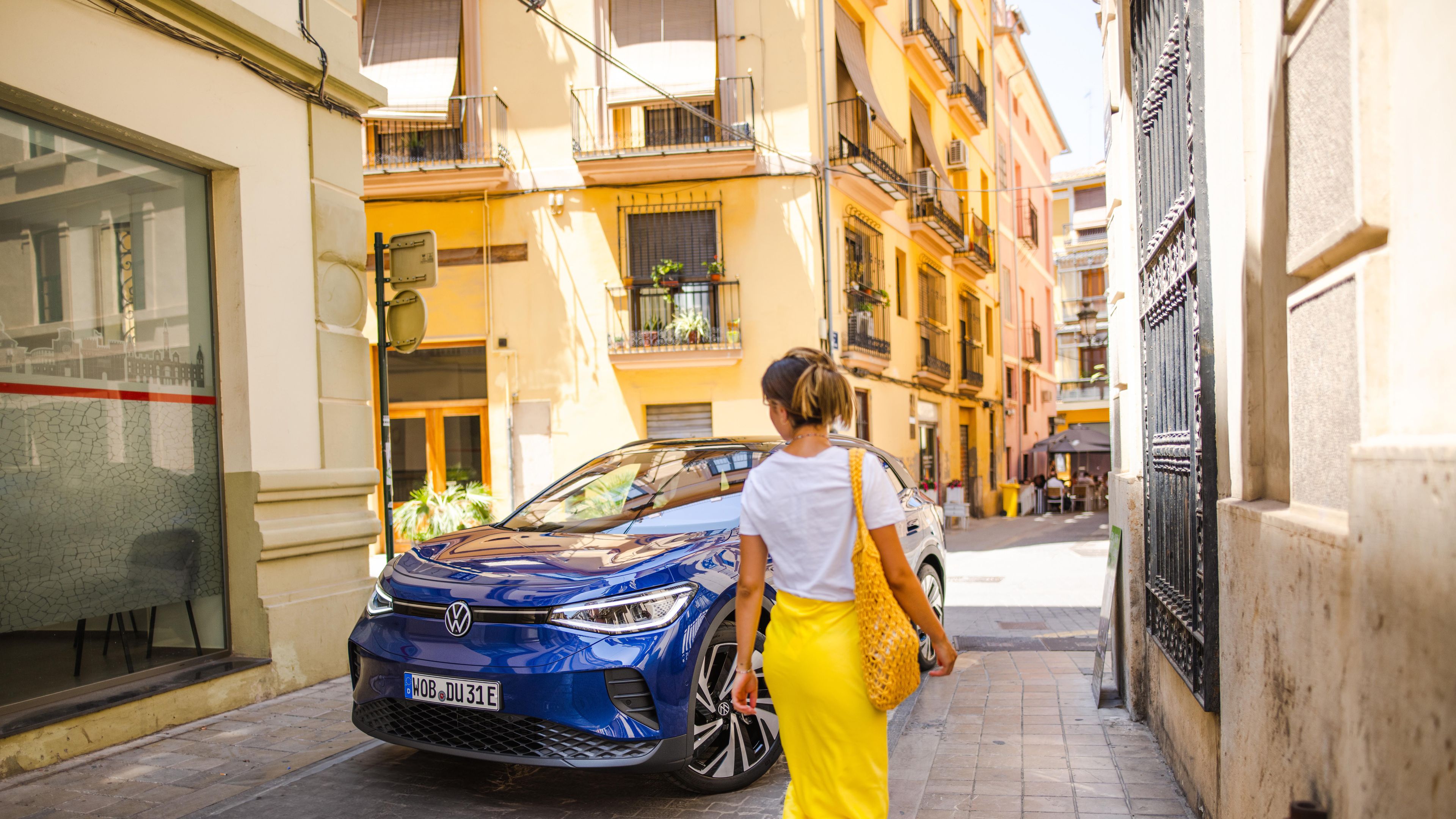 A lady in a yellow skirt walks up to the Volkswagen ID.4 in the colour Blue Dusk Metallic in a narrow walkway surrounded by tall buildings.