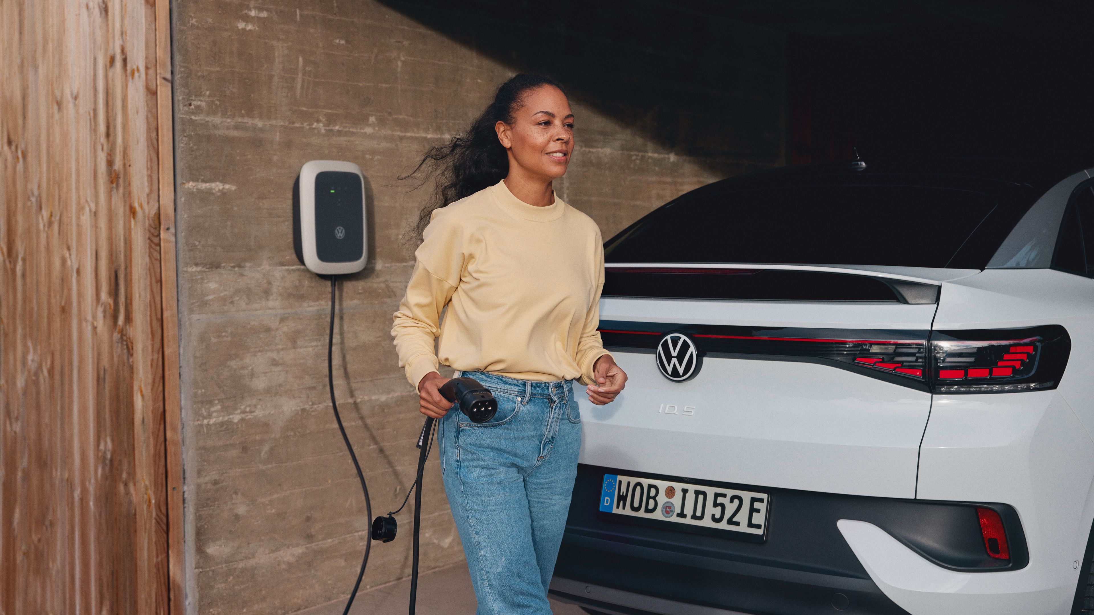 A lady wearing yellow blouse holding the battery charging plug while walking toward the White Volkswagen ID.5 