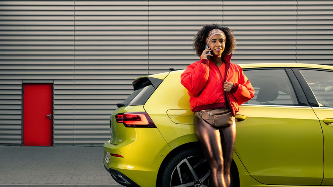 A chic looking lady making a phone call while leaning against a yellow Volkswagen Golf parked in front of a building.