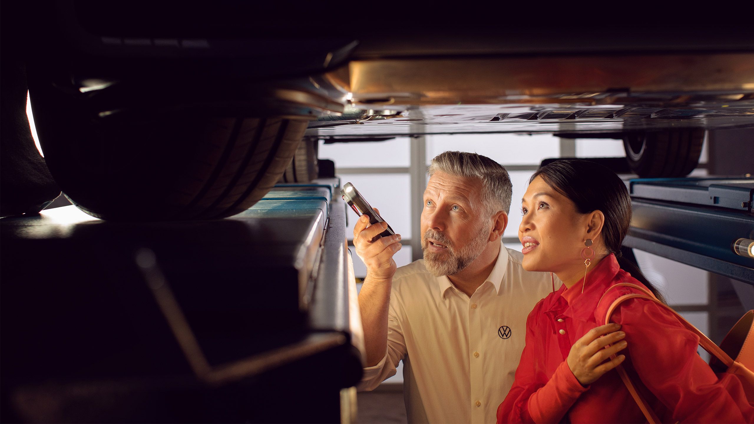 A Certified Volkswagen Staff using a torchlight to direct the attention a female customer dressed in red to the undercarriage of her Volkswagen vehicle in a service workshop.