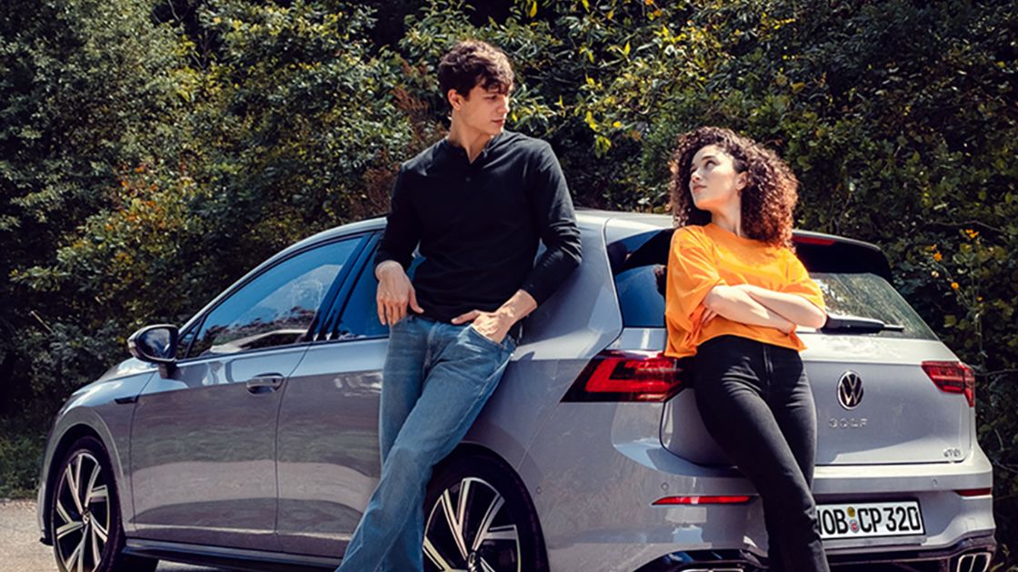 Two young individuals in casual outfits looking at each other while leaning against a silver coloured Volkswagen Golf at an outdoor setting.