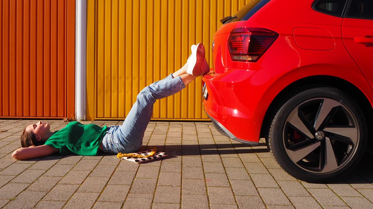 A casually dressed lady lying down and resting her legs on the bumper of a red Volkswagen vehicle parked beside an outdoor urban area under a sunny weather.