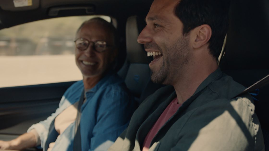 A young adult driving a Volkswagen vehicle is seen having an enjoyable conversation with an old individual seated in the passenger seat.