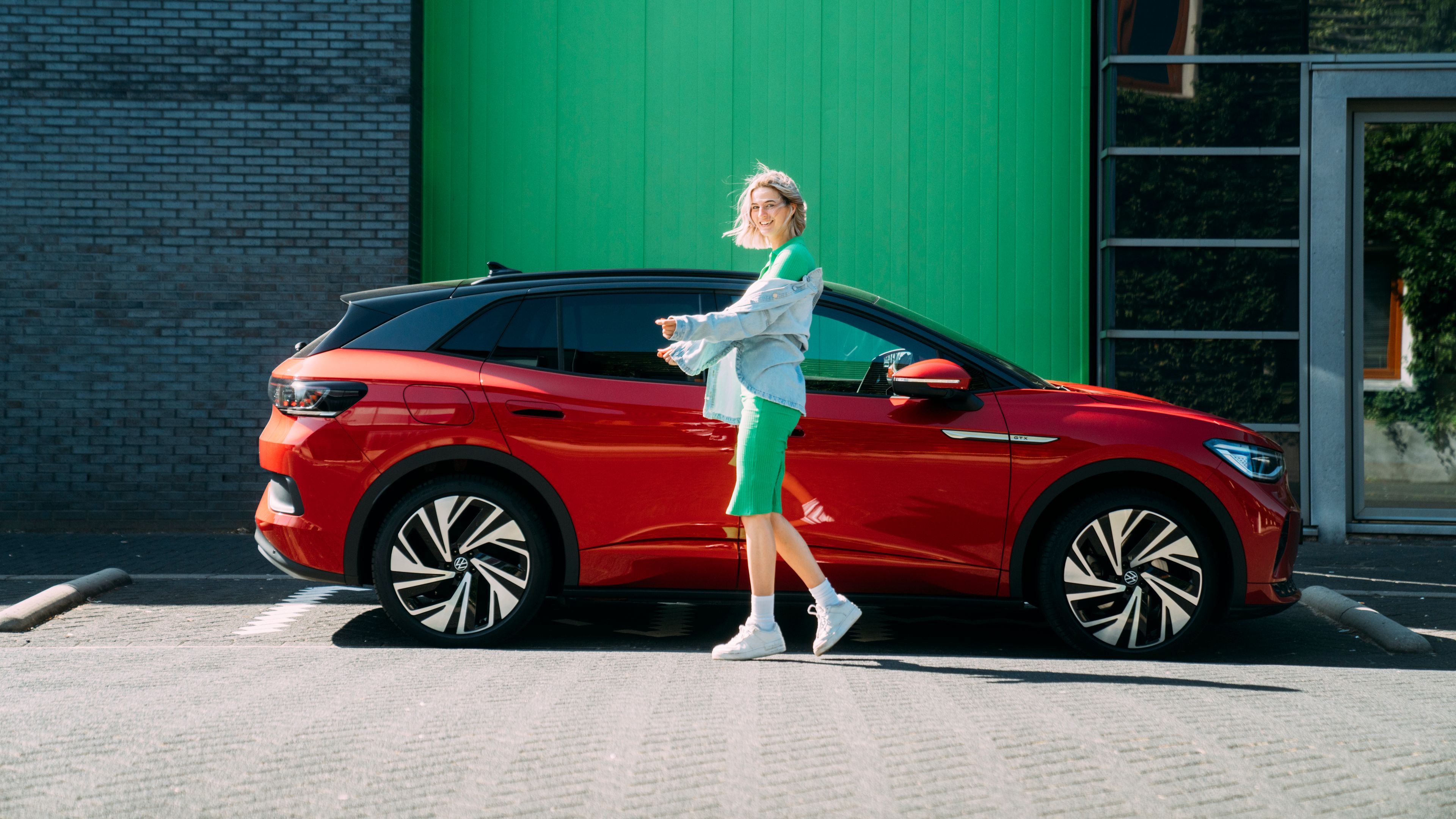 A lady in a green dress walks up to the Volkswagen ID.4 GTX in the colour Kings Red Metallic in a front of a largebuildings.