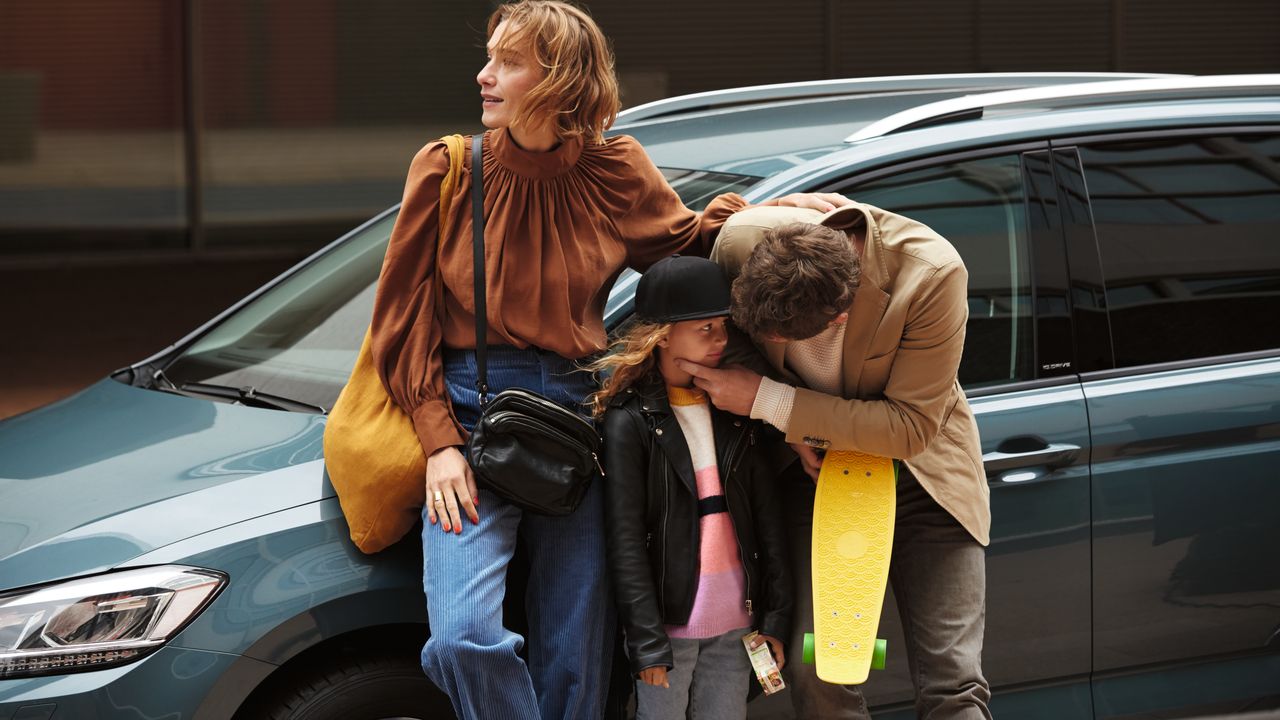 A family standing beside of a parked Volkswagen vehicle while a parent is checking the condition of their child up-close at an outdoor urban area.