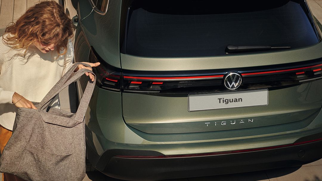 A lady is seen walking away from a metallic green Volkswagen Tiguan while fiddling with her tote bag at day.